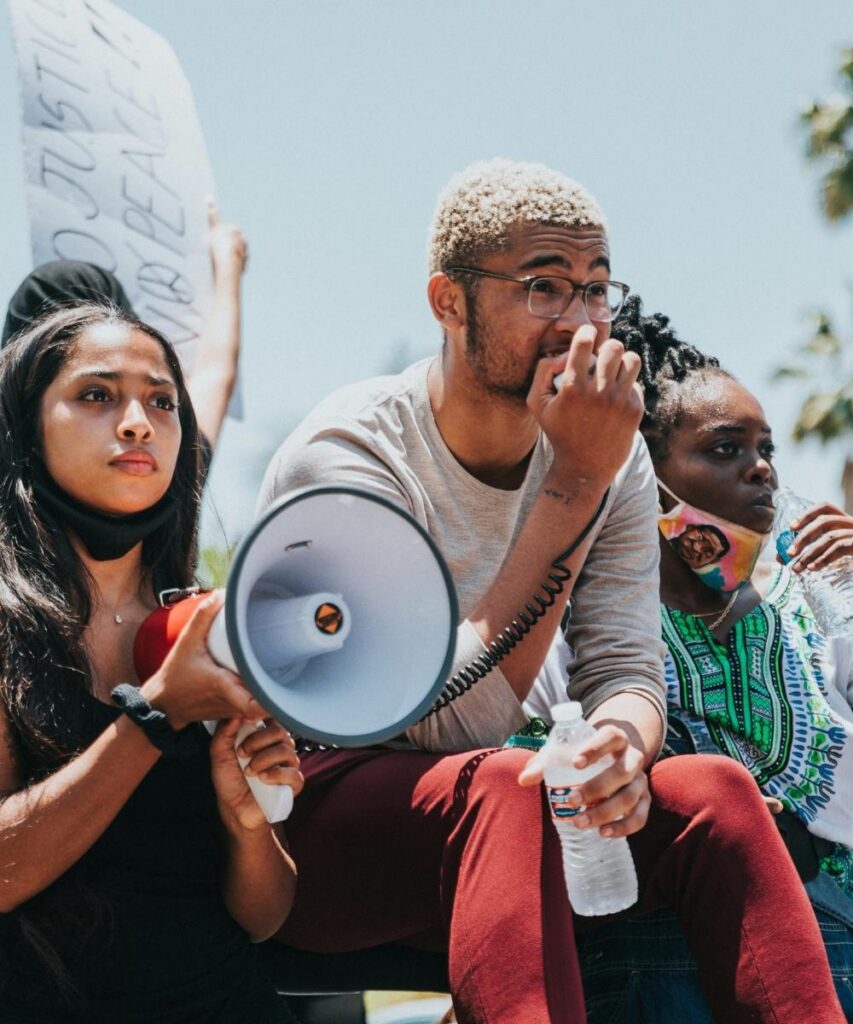 A medium shot of three young activists at an outdoor protest; a young woman on the left holds a white megaphone and looks off-camera with a serious expression while a cardboard protest sign reading 'JUSTICE... PEACE' is visible behind her, a young man with short blonde hair and glasses sits in the center holding a water bottle, and another young woman wearing a patterned top stands on the right side of the group—Team DSB Young People Address the Nation case study rally asset